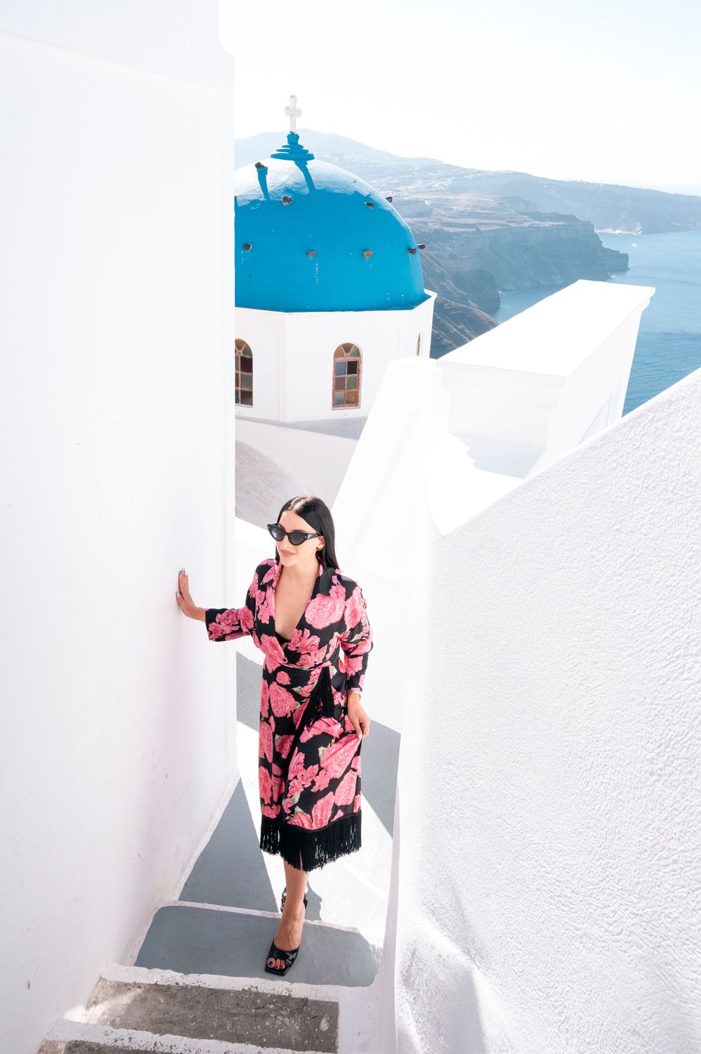Woman in pink floral dress poses on white steps with iconic blue domed church in Santorini, Greece.