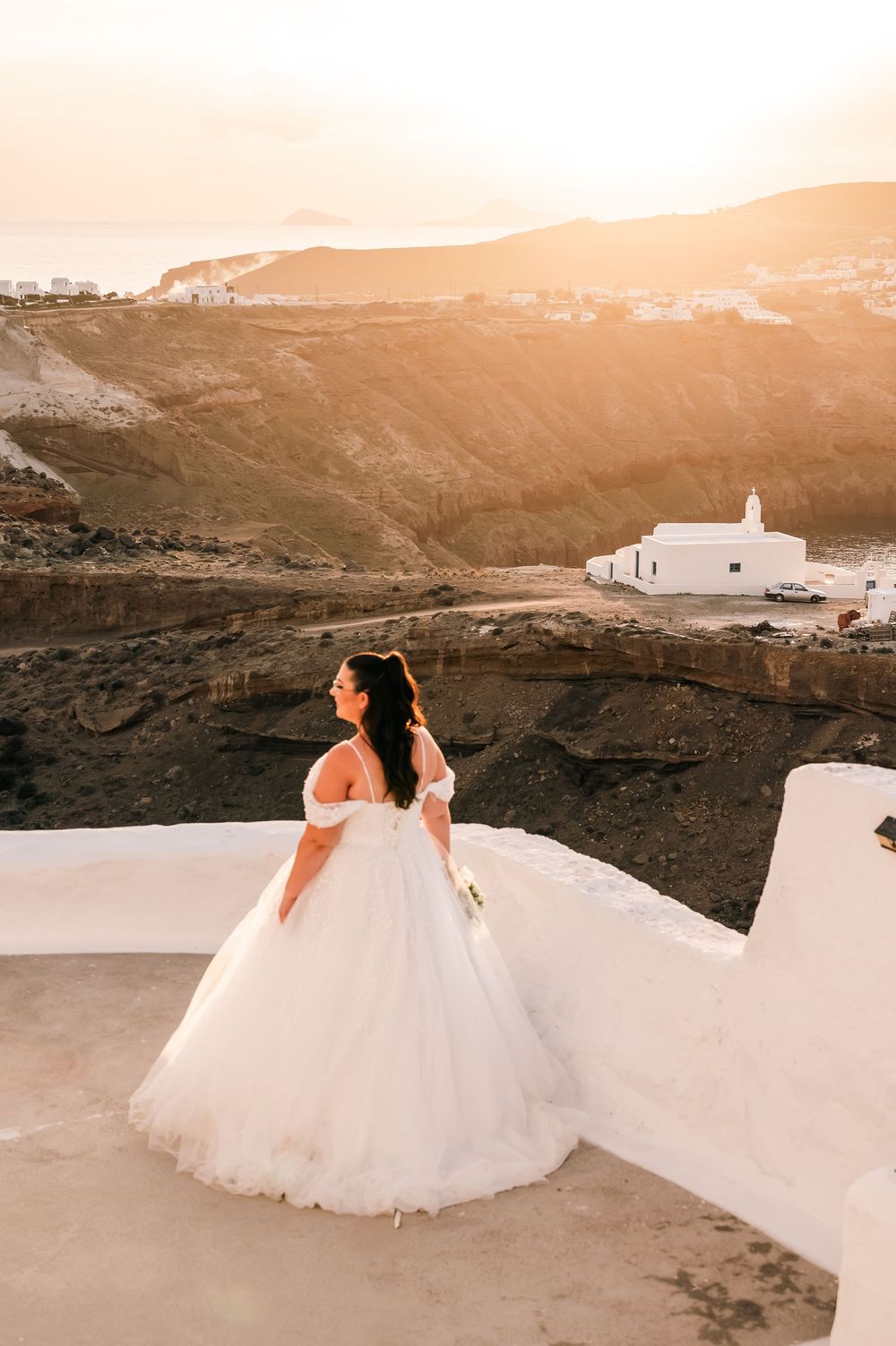 Bride in elegant ball gown poses on white terrace at golden sunset with Santorini landscape behind her.