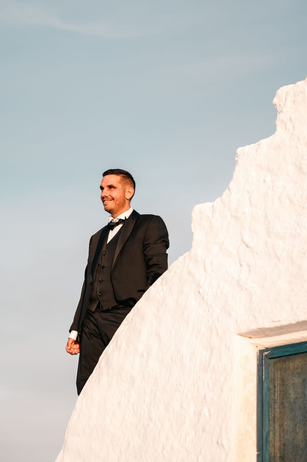 Elegant groom in black tuxedo stands confidently beside whitewashed Greek architecture under blue sky.