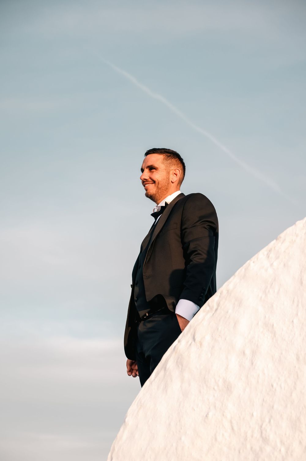 Smiling groom in black tuxedo and bow tie standing on white structure against blue sky.