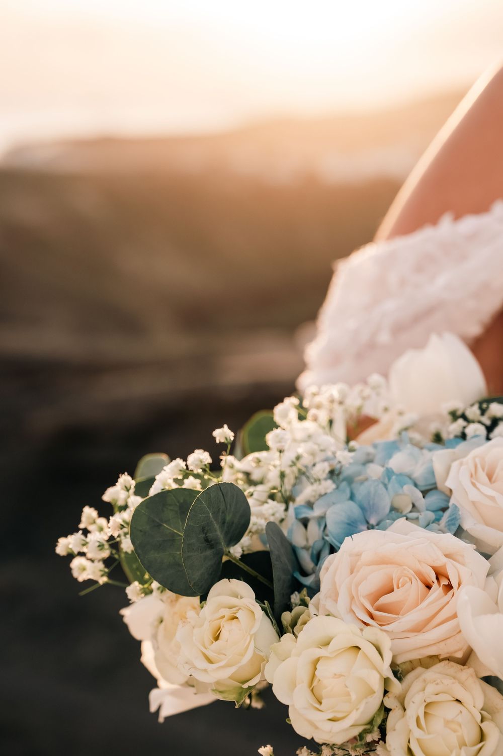 Close-up of a bridal bouquet with white roses, blue hydrangeas, and eucalyptus leaves in golden sunset light.