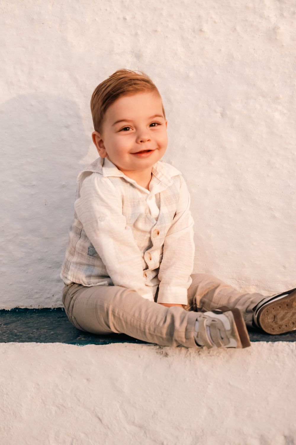Smiling toddler boy in plaid shirt and khaki pants sitting against a white wall in warm sunlight.