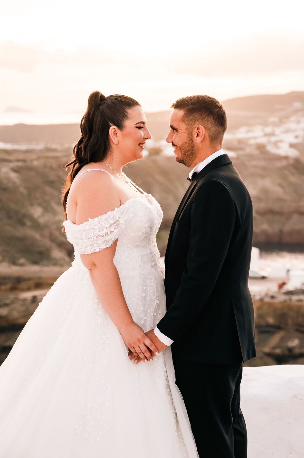Bride in off-shoulder white ball gown and groom in black tuxedo share a romantic moment at sunset overlooking the ocean.