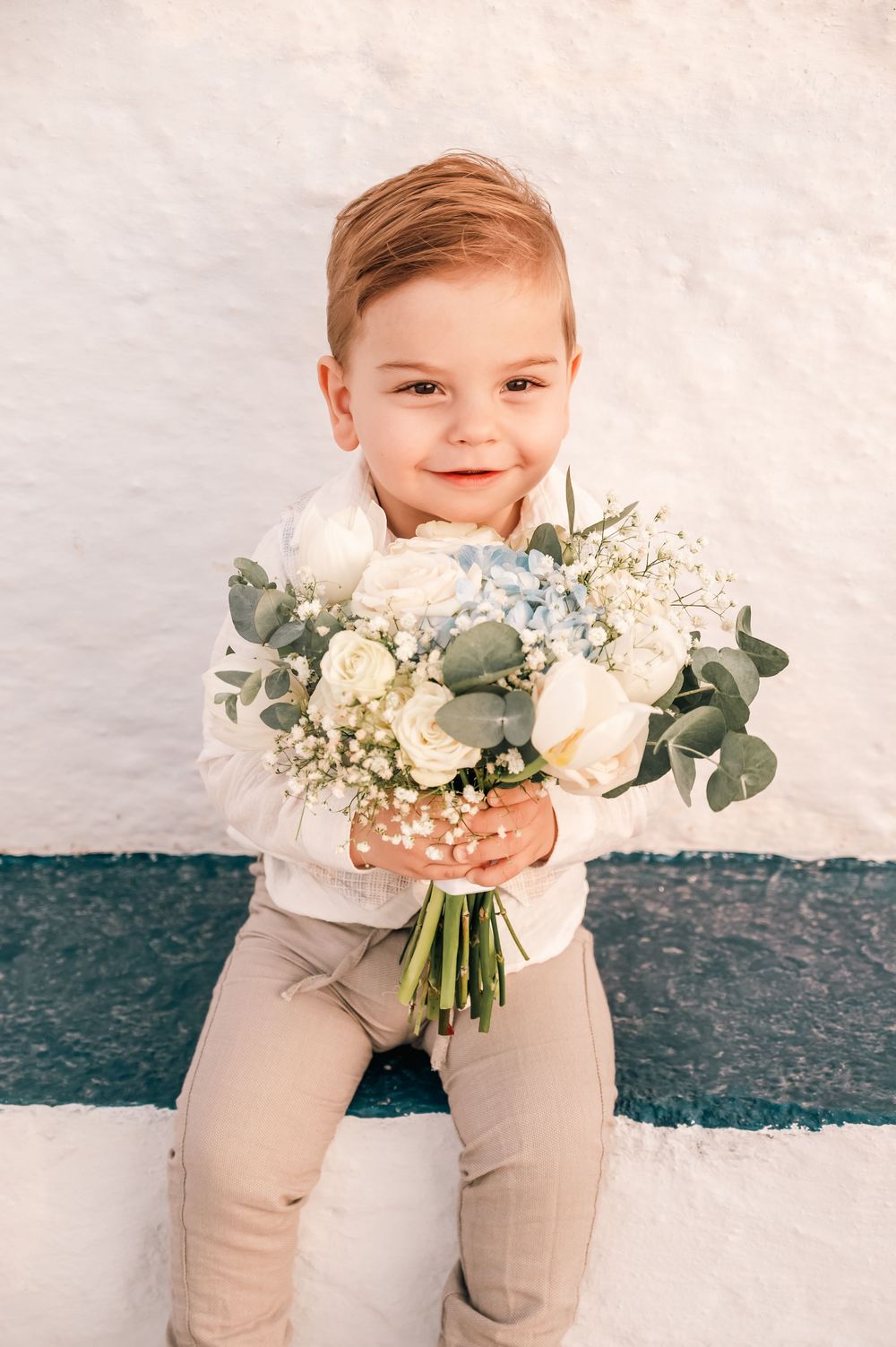 Smiling toddler boy holding a white and blue wedding bouquet with roses and eucalyptus while sitting outdoors.