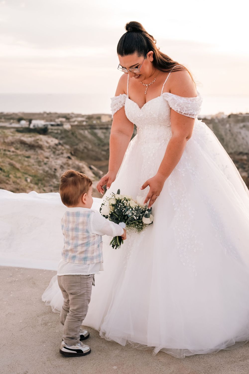 Bride in off-shoulder ball gown holding bouquet while interacting with young toddler boy at sunset wedding.