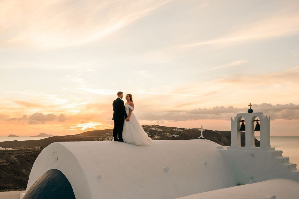 Bride and groom share a romantic sunset kiss atop a white chapel in Santorini, Greece.