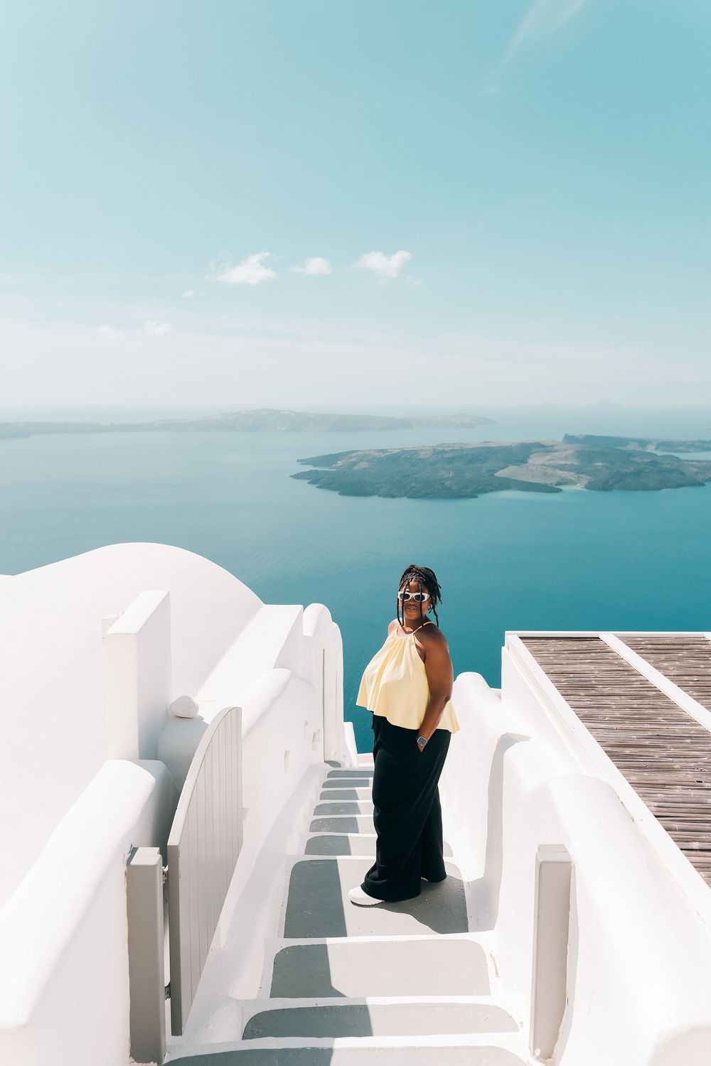 Woman in yellow top and black pants poses on white Santorini steps with stunning Aegean Sea and volcanic island backdrop.