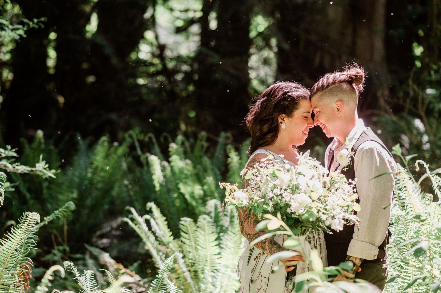 two brides rest forehead to forehead in a lush garden setting surrounded by white flowers and greenery.