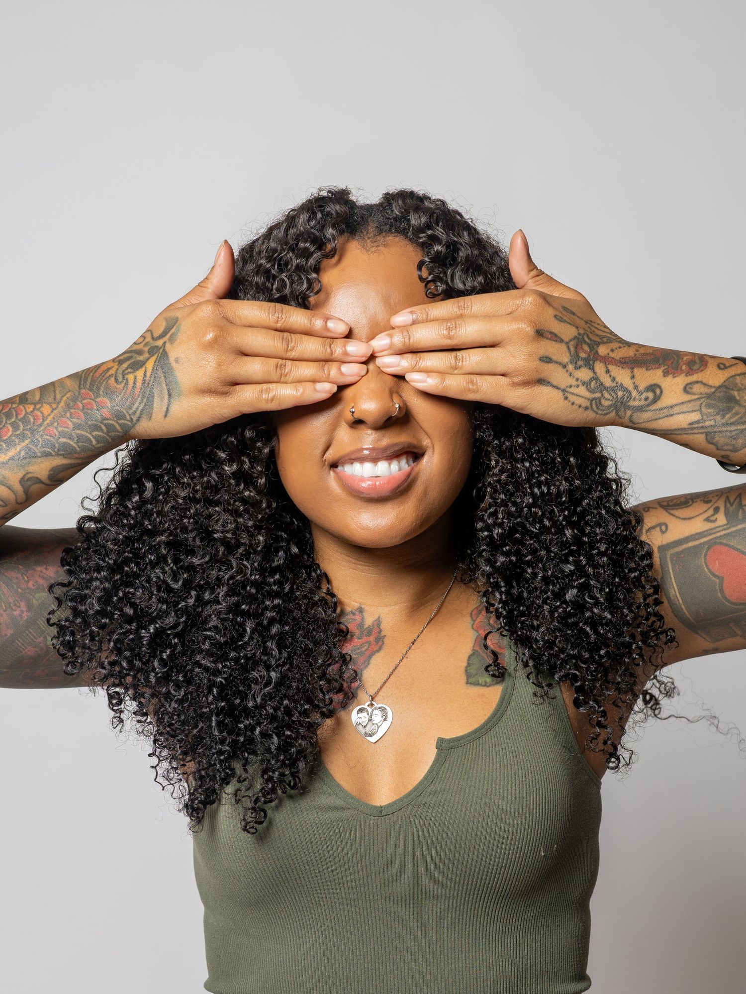 Tattooed woman with curly hair covers her eyes with both hands and smiles playfully.