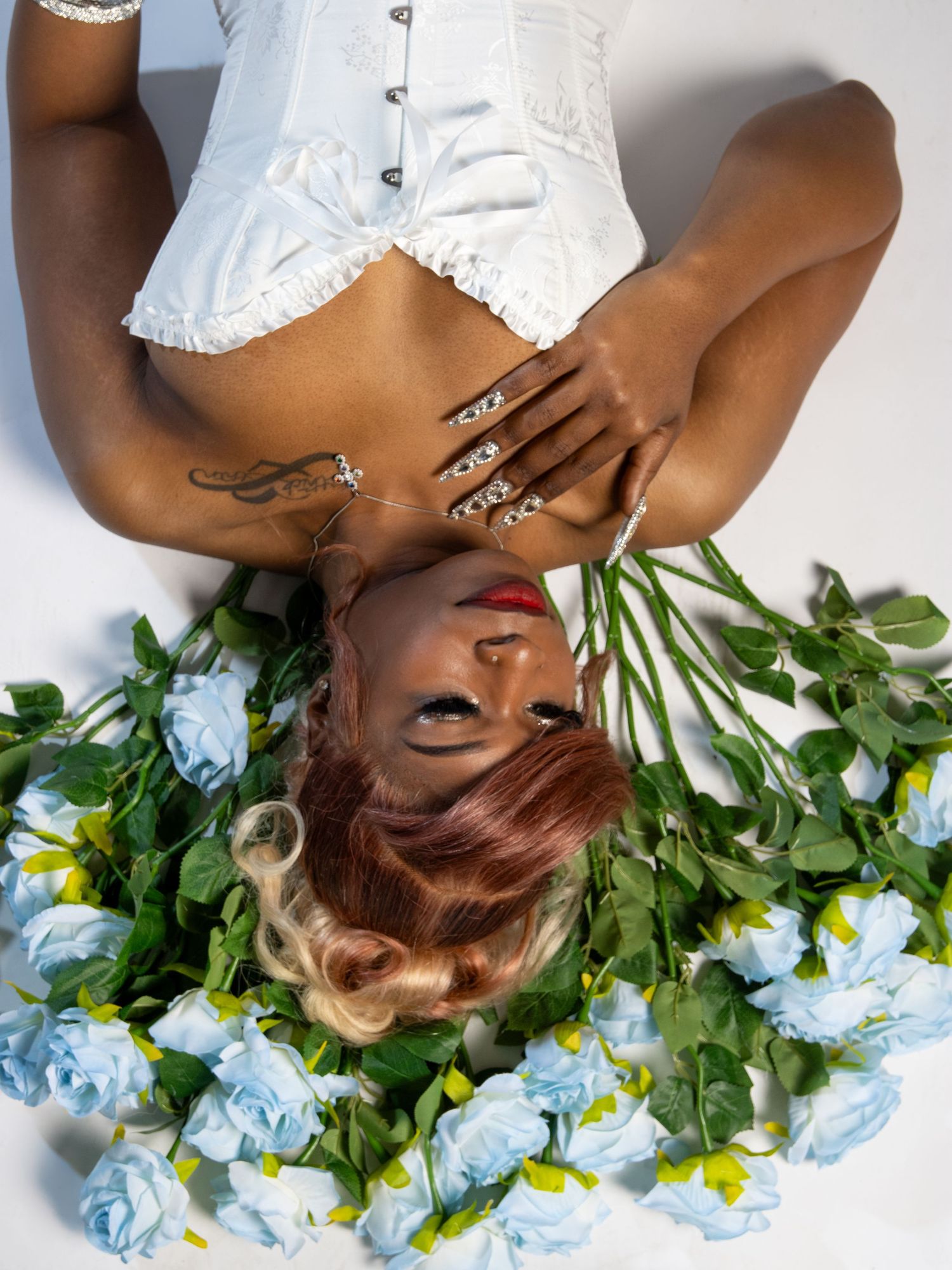 Woman in white corset lies among blue roses in studio.