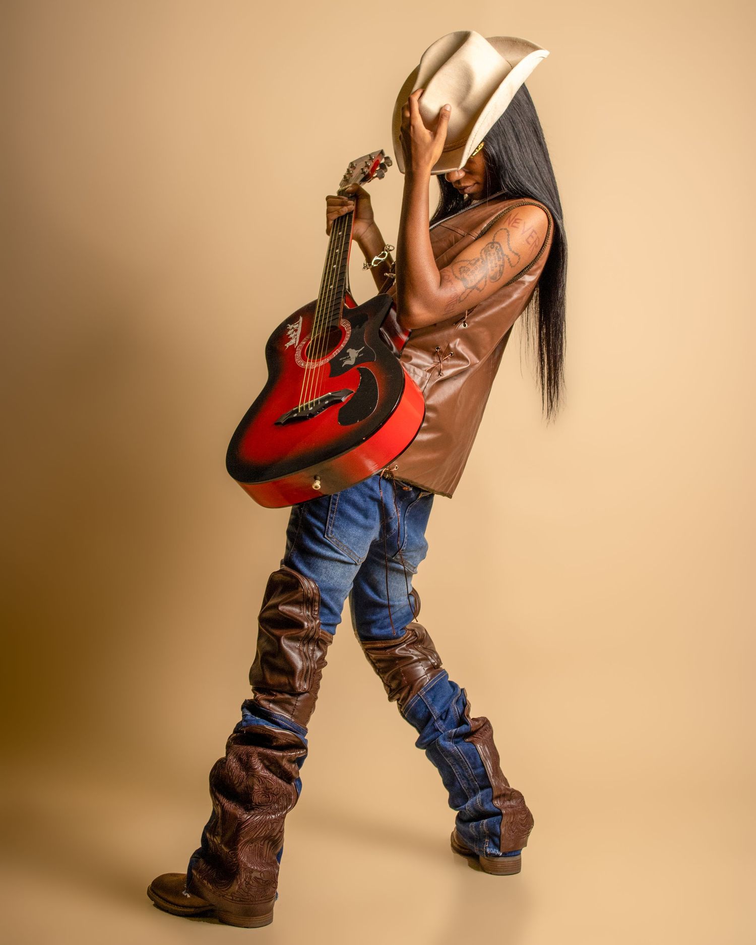 A musician poses with a red acoustic guitar and white cowboy hat, wearing jeans and brown boots.