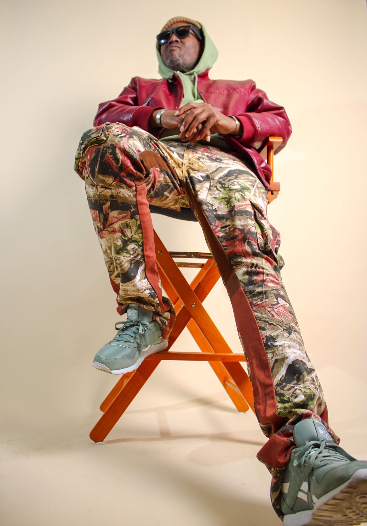 Stylish man in colorful pants and red jacket poses on orange stool with sunglasses.