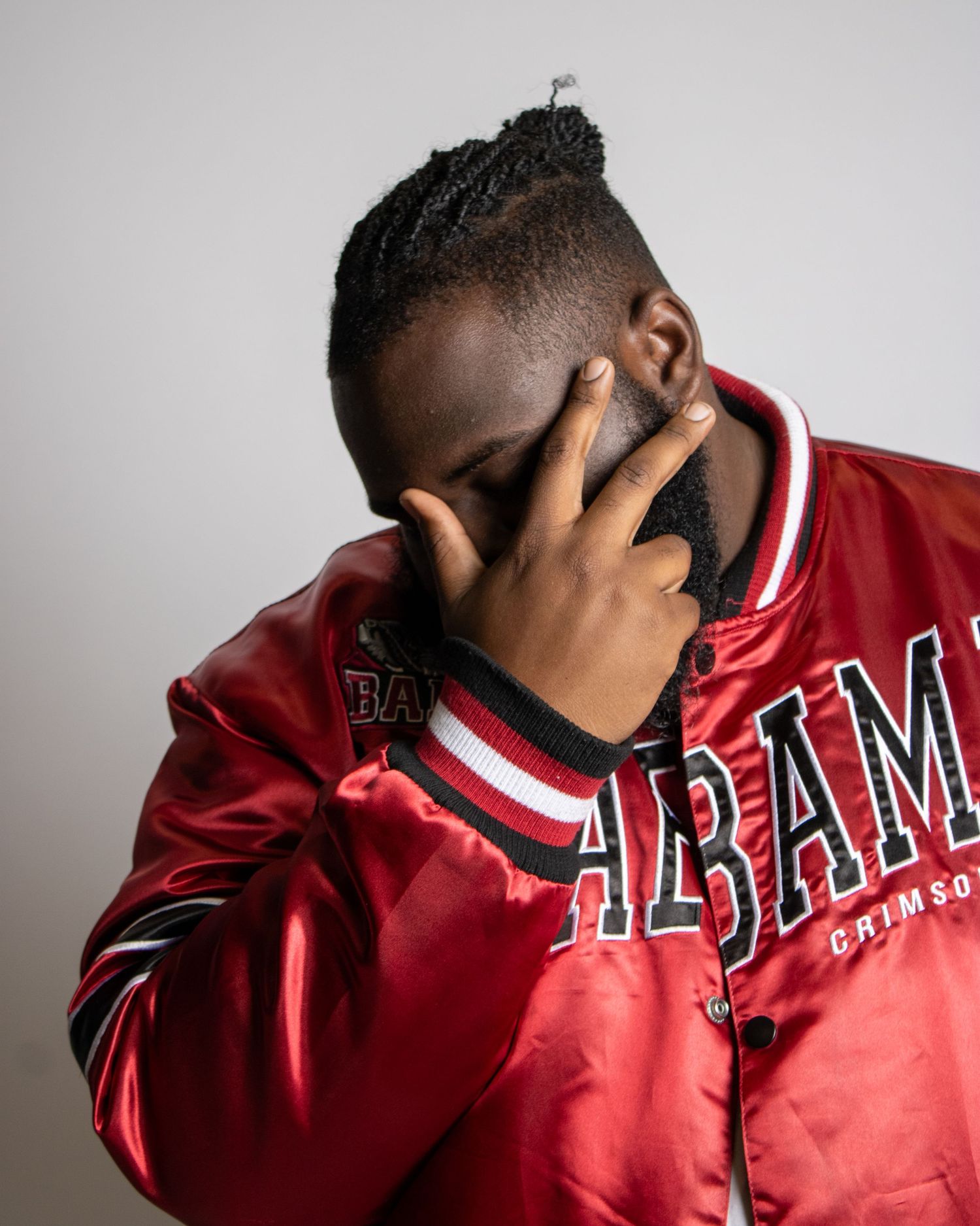 Man in red Alabama Crimson Tide varsity jacket covers face with hand in portrait.