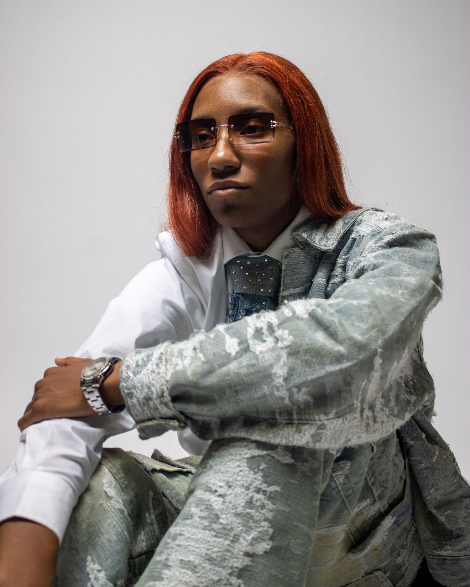 Person with red hair wearing stylish silver jacket and sunglasses in artistic pose.