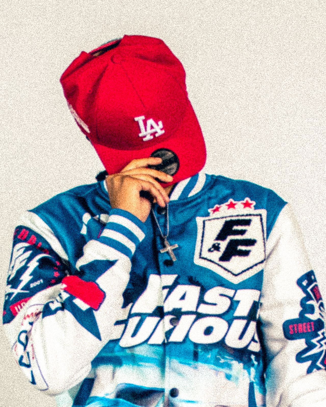 Person in Fast and Furious jacket wearing red LA cap holding microphone outdoors.