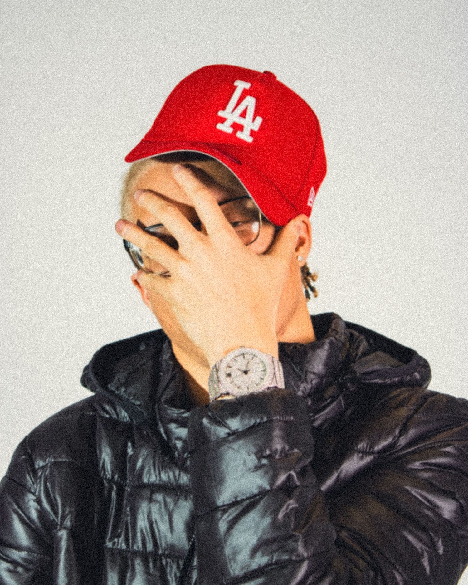 Person in red LA cap and black puffer jacket covering face showing tattooed wrist.