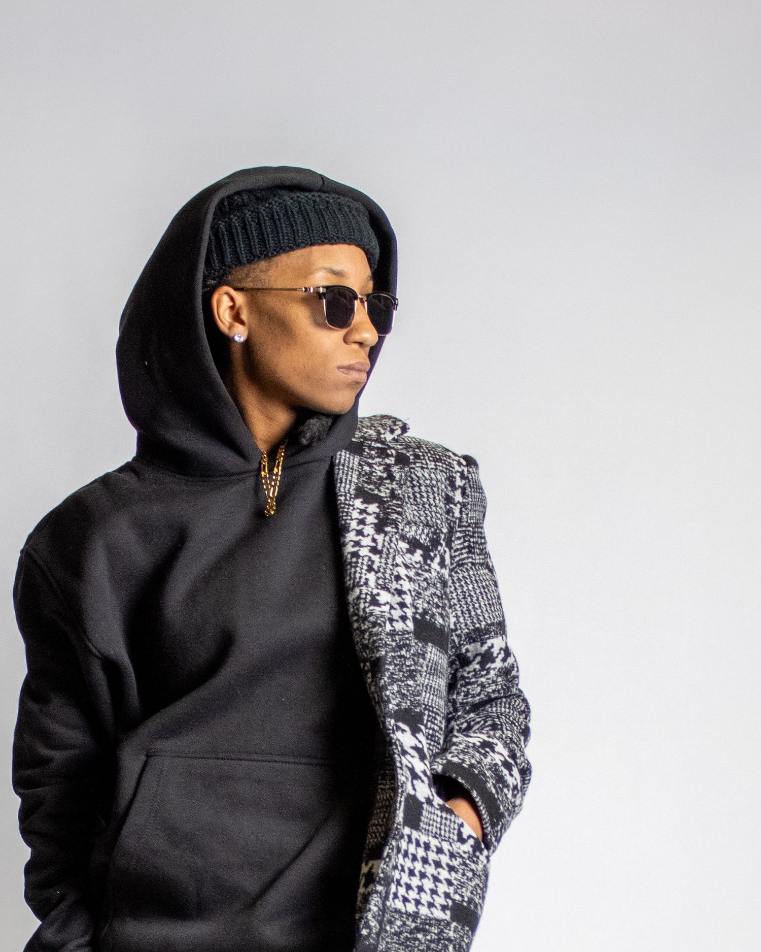 Stylish man in black hoodie and patterned coat wearing sunglasses and beanie.