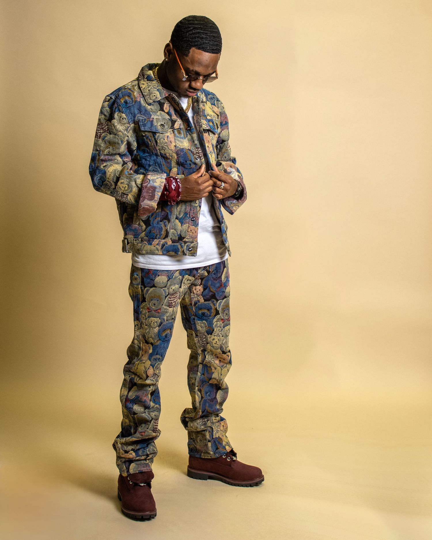 Man posing in matching floral tapestry jacket and pants with burgundy boots on gold background.