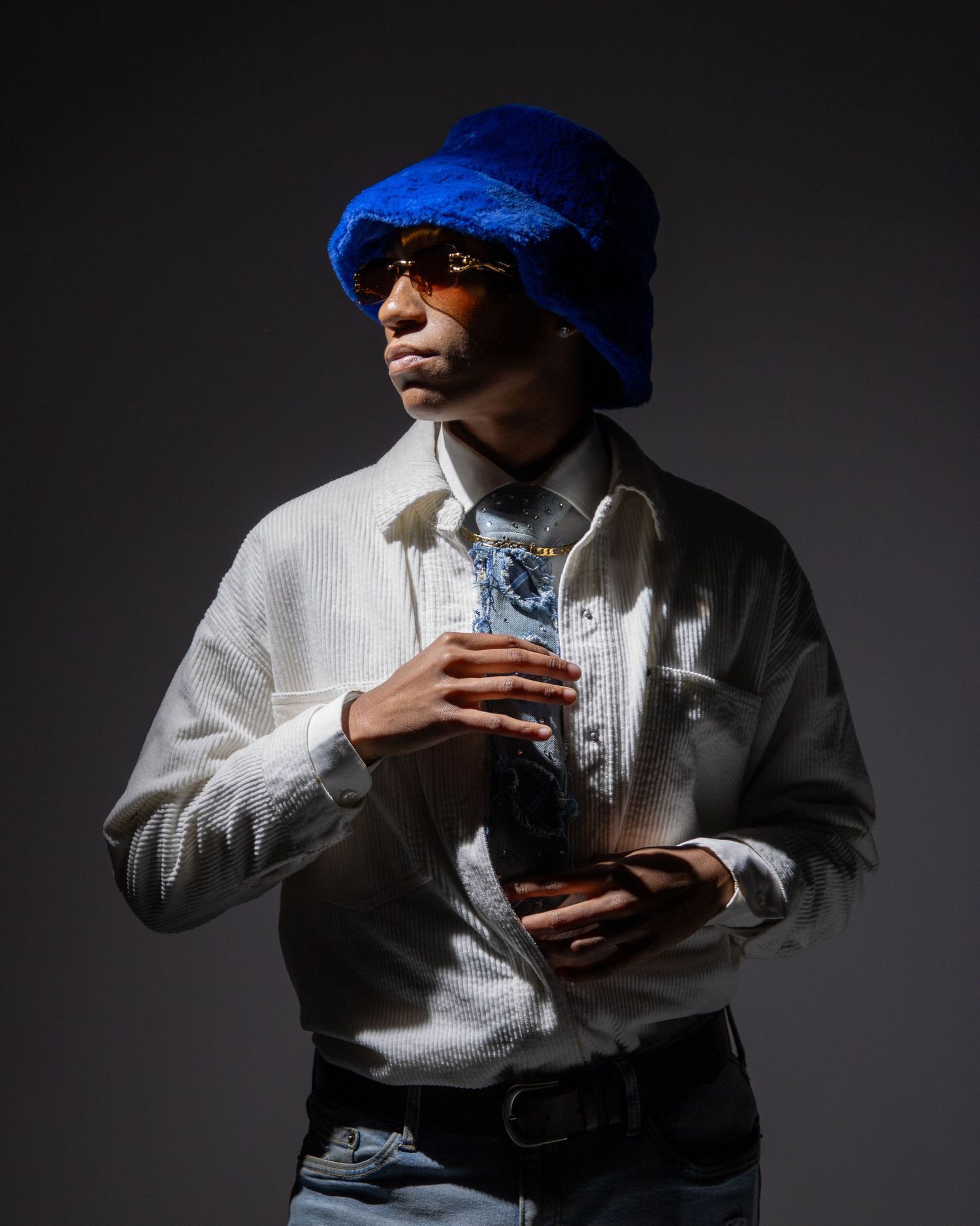 Stylish man wearing blue bucket hat, white shirt, and sunglasses holding a drink in dark studio.