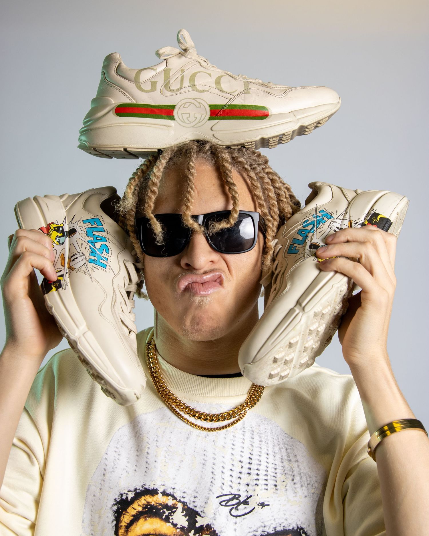 Person holding Gucci sneakers beside their head wearing sunglasses and gold chain necklace.