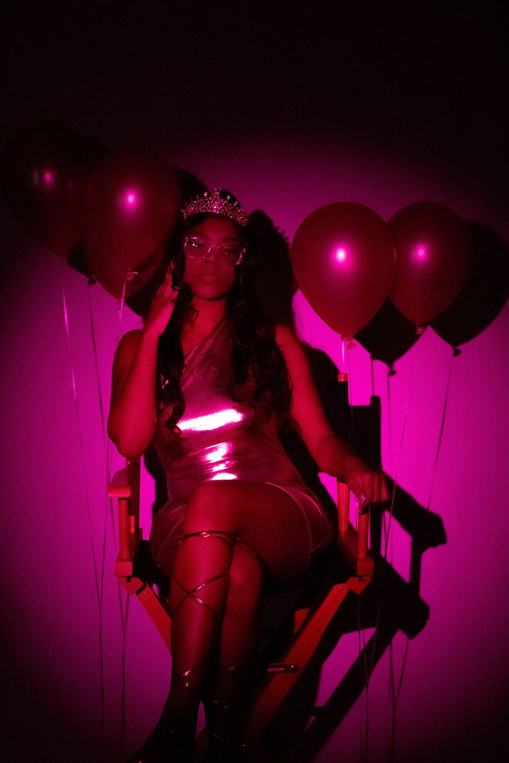 Woman wearing tiara sits in director chair surrounded by pink balloons in magenta lighting.