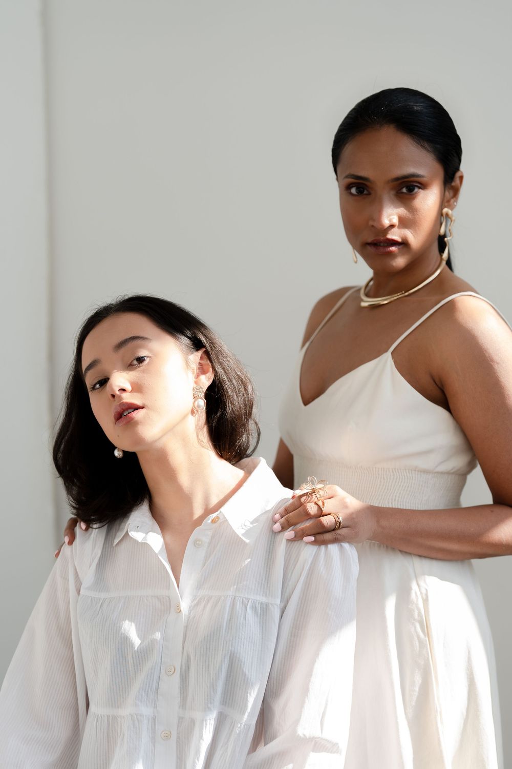 Two models in contrasting white outfits pose artistically against a neutral wall in minimalist fashion photoshoot.