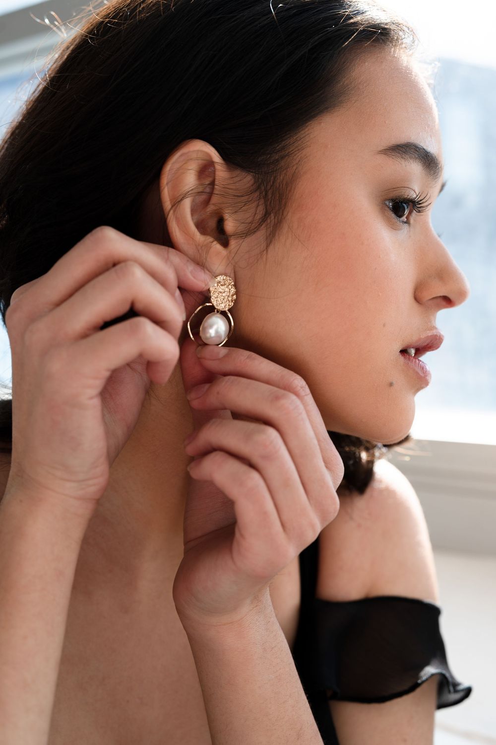 Someone putting on an elegant pearl and gold statement earring in front of a window with natural lighting.