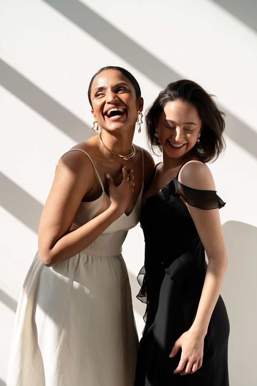 Two friends in contrasting white and black dresses share a joyful candid moment against a light background.