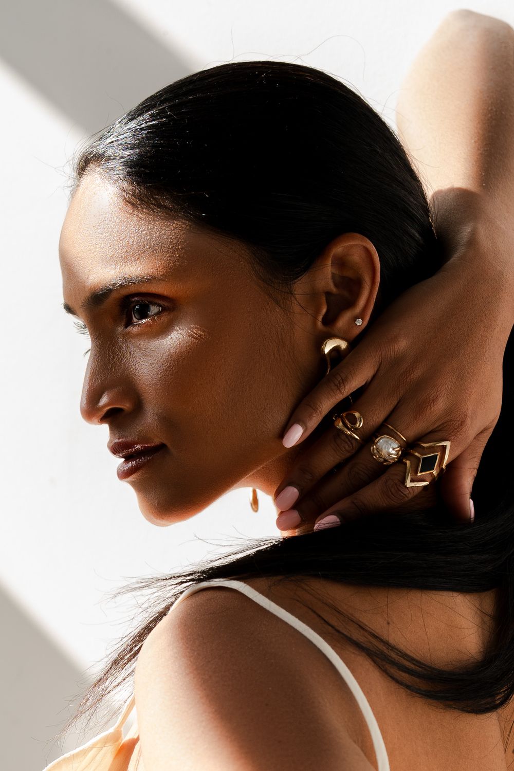 Woman with dark hair wearing white top and statement geometric rings poses in dramatic side lighting.