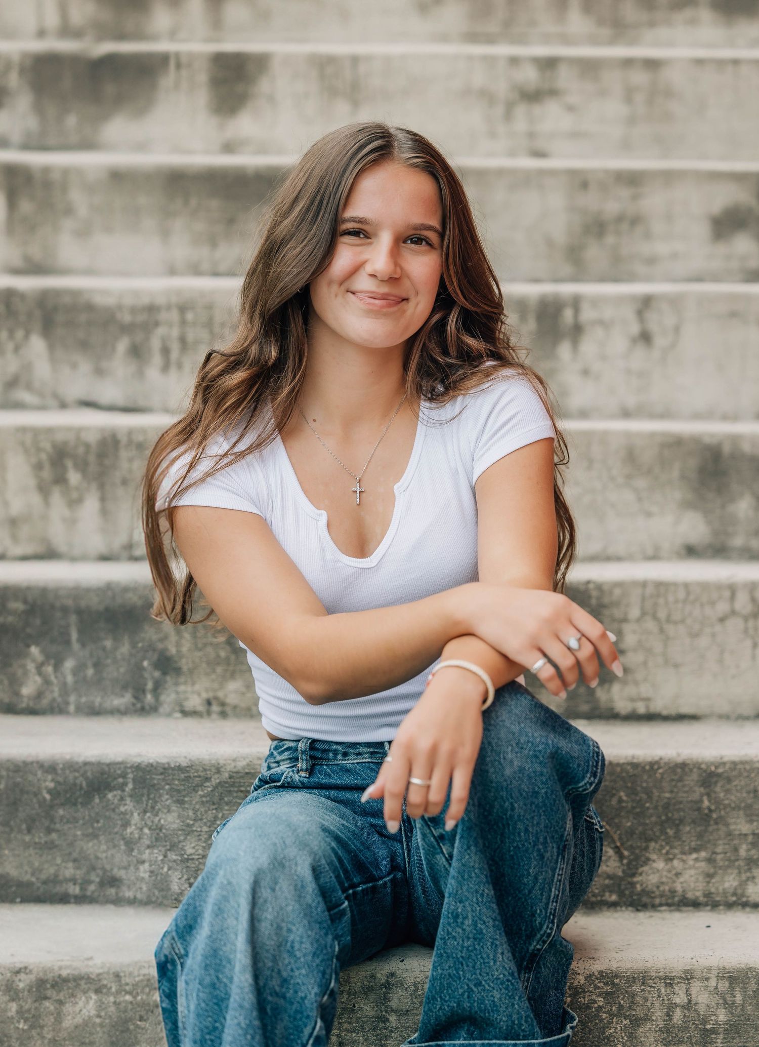 Young woman with long brown hair sits on stone steps, wearing white tee and jeans, smiling warmly.