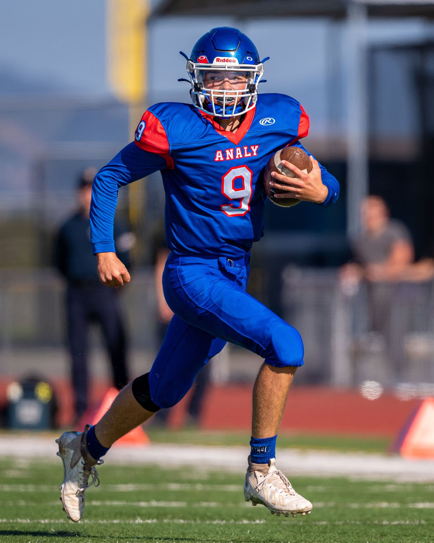 Football player in blue and red uniform carries ball while running downfield.
