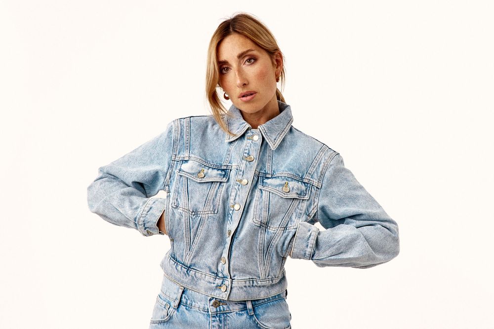 Woman in light blue denim jacket and jeans posing with hands on hips against white background.