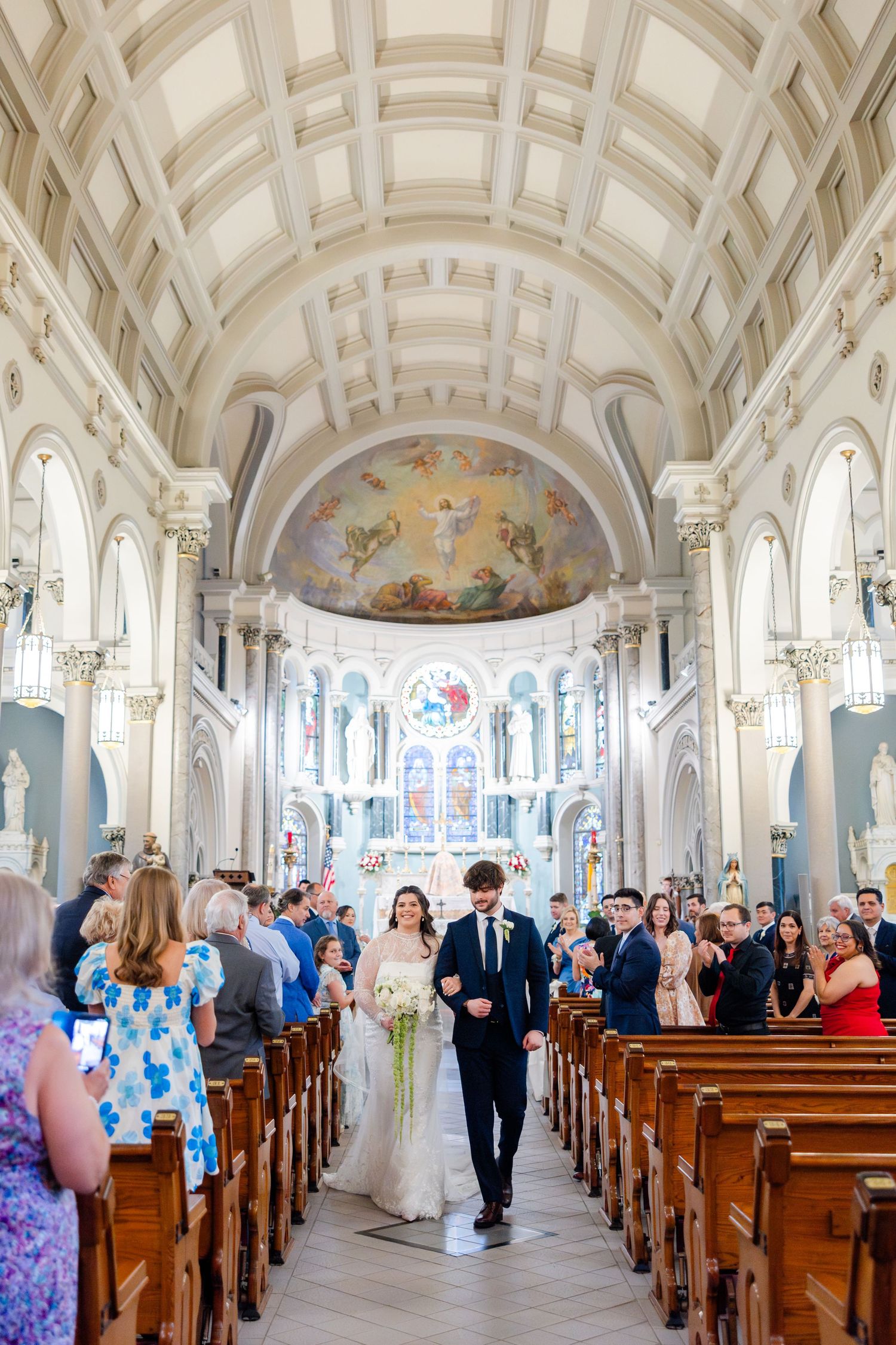 Wedding ceremony in a beautiful white cathedral with arched ceilings and stained glass windows as guests fill the pews.