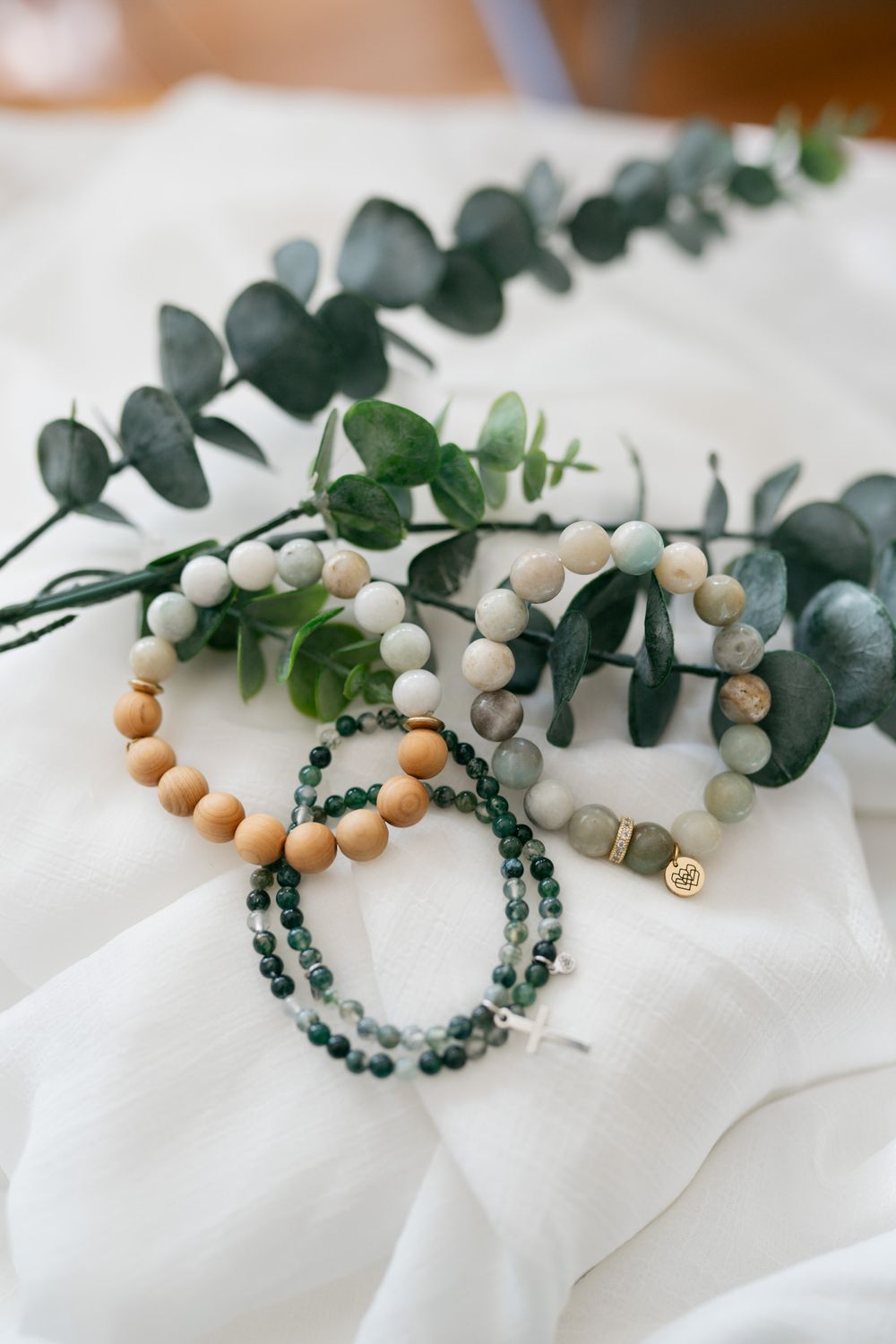Beaded bracelets with wooden and white beads displayed with fresh eucalyptus sprigs on white fabric.
