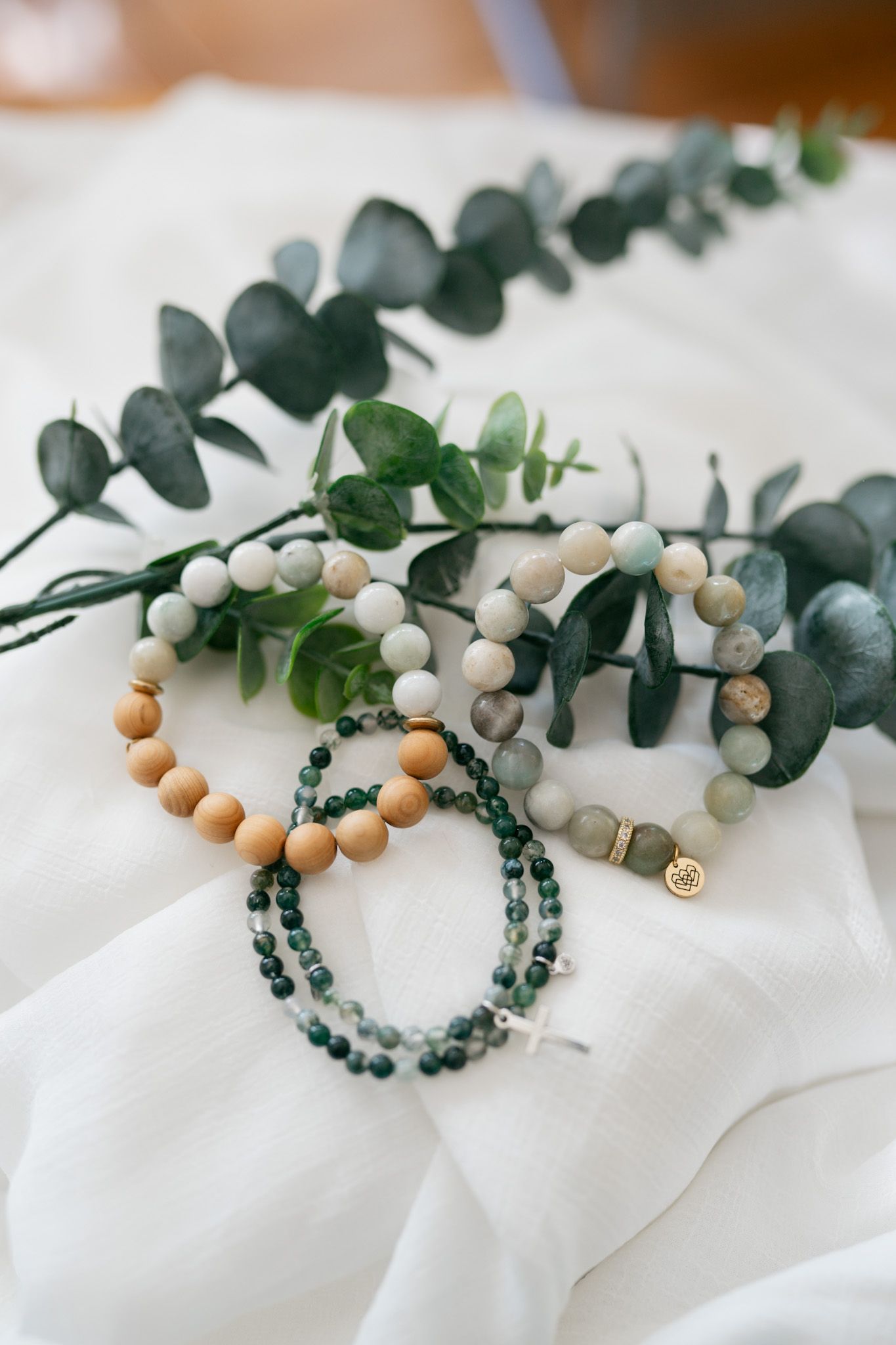 Beaded bracelets with wooden and white beads displayed with fresh eucalyptus sprigs on white fabric.