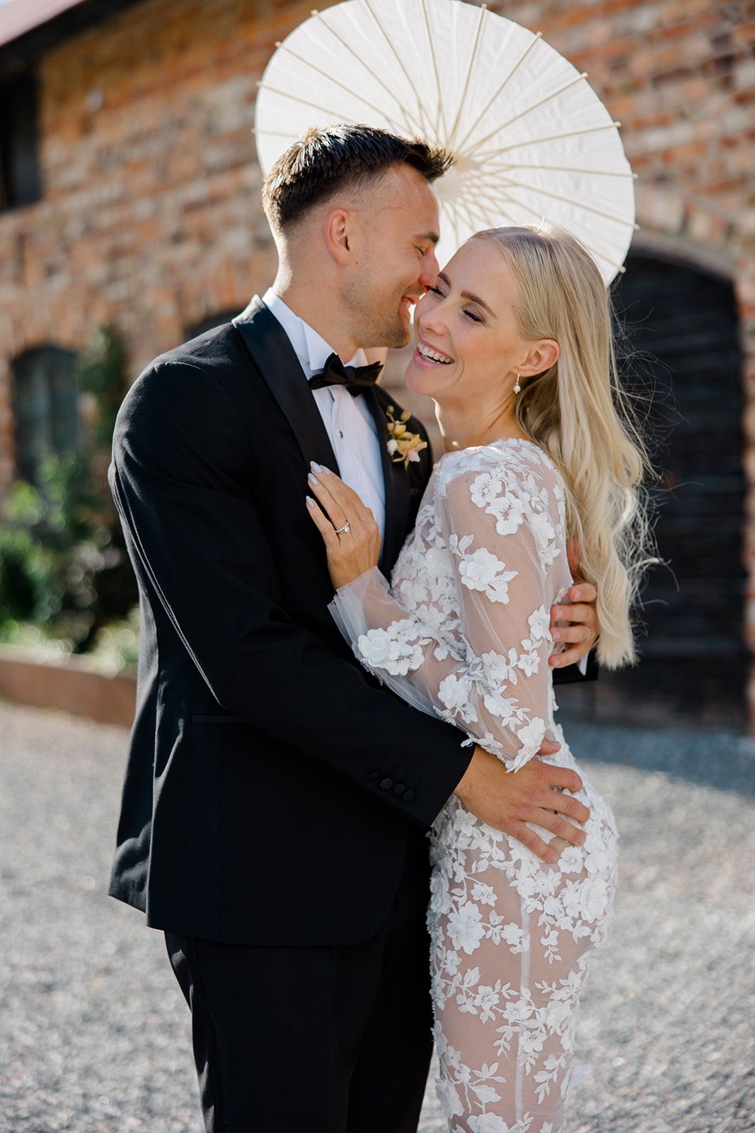 Newlywed couple shares intimate moment under white parasol, bride wearing sheer lace gown and groom in classic tuxedo.