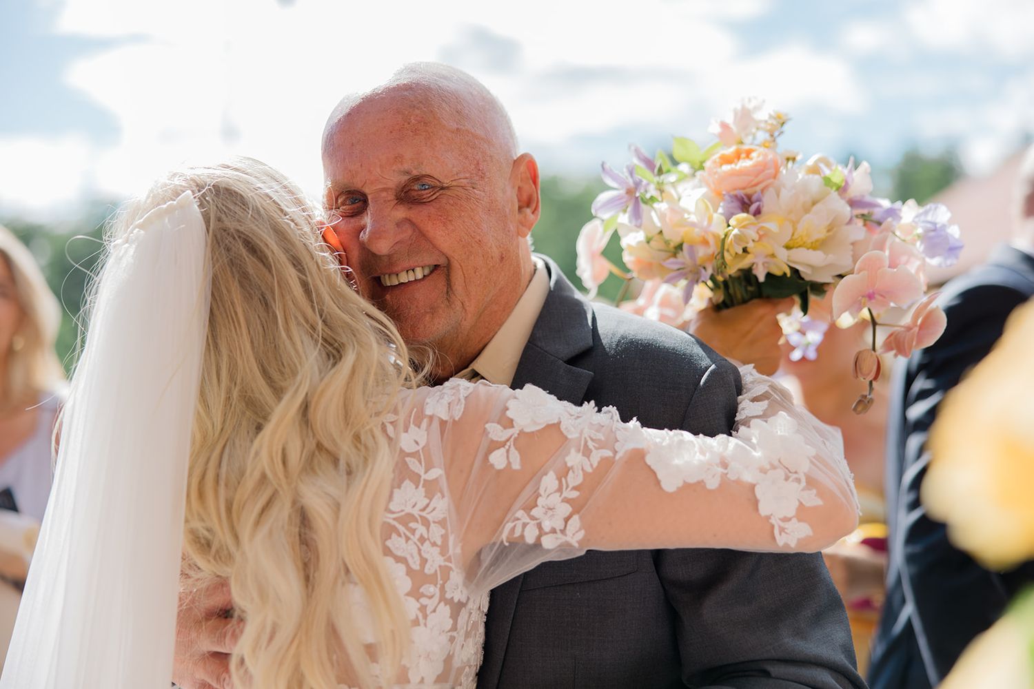 A couple in wedding attire shares a romantic kiss under a wooden pergola decorated with pink and white flowers.