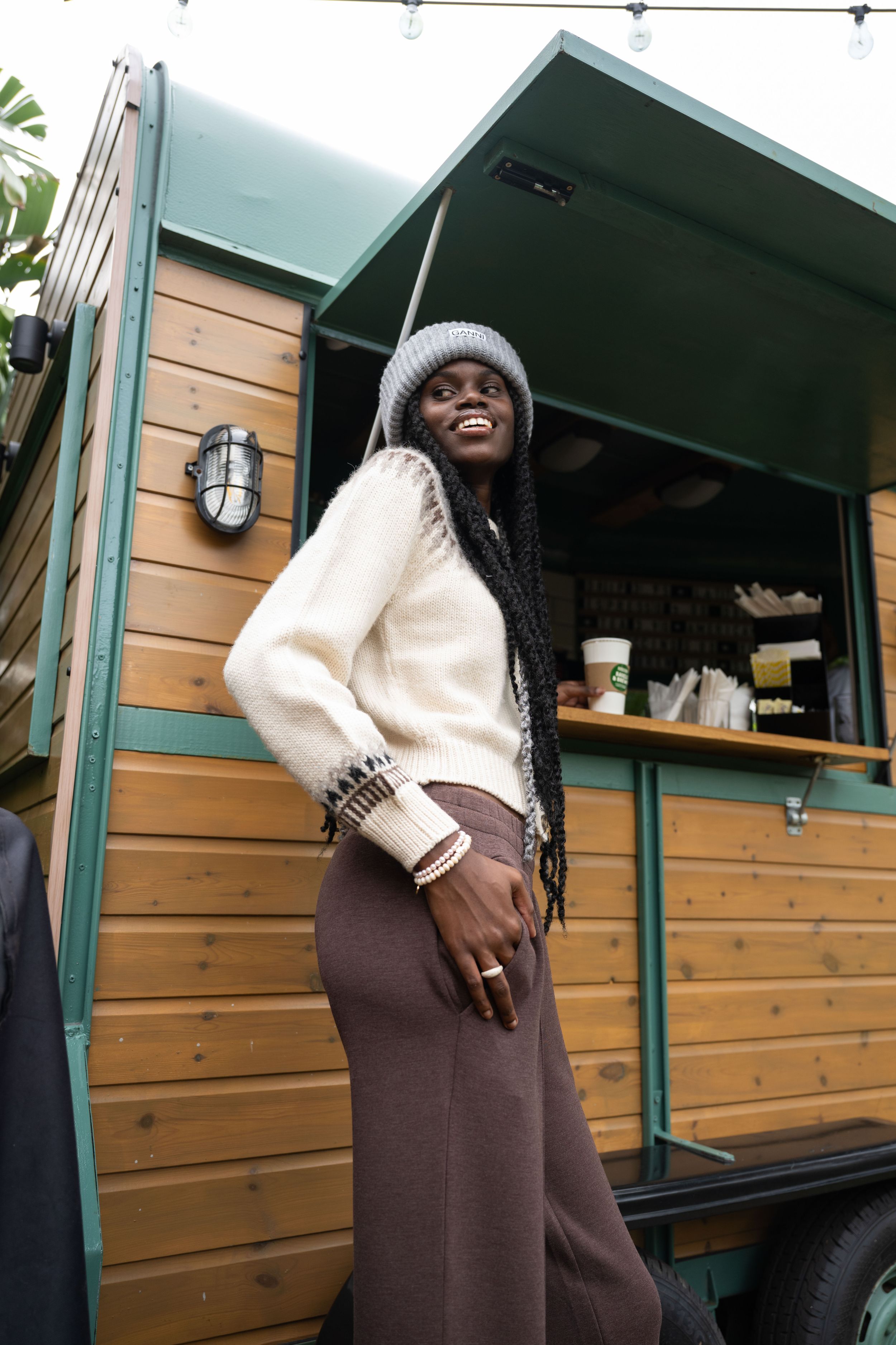 A person wearing a white sweater and brown skirt stands smiling by a wooden cabin exterior with green trim.