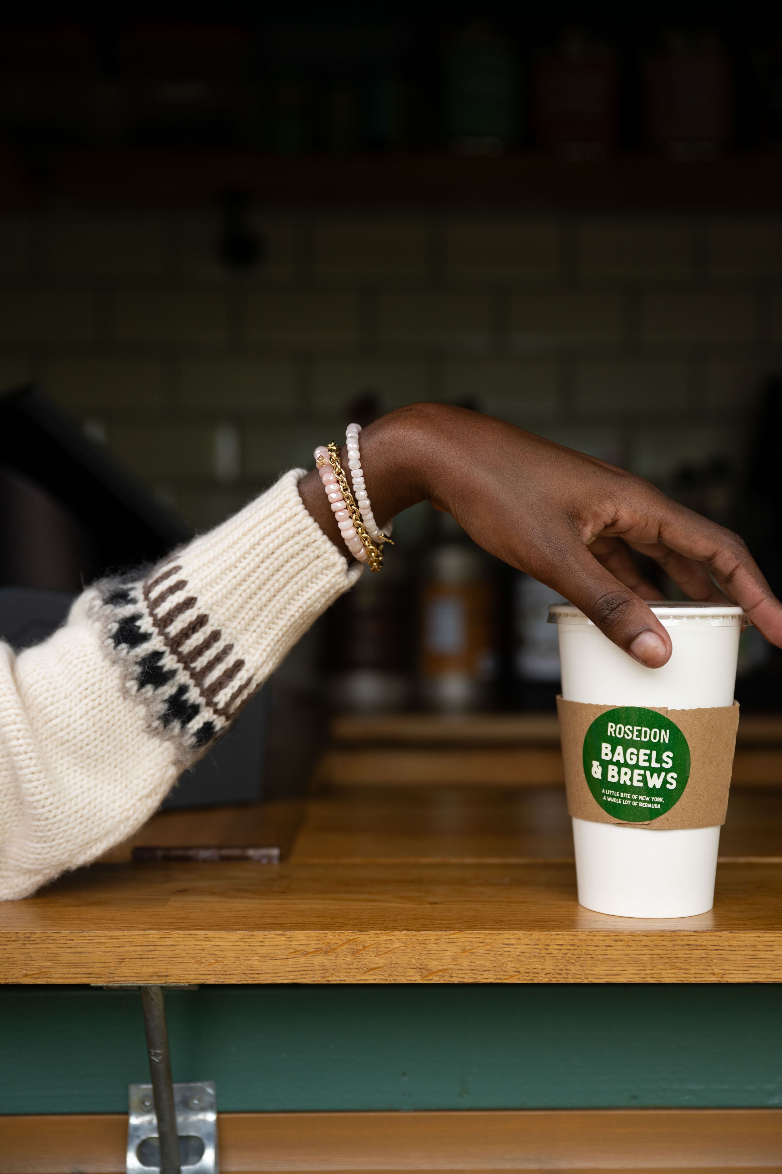 Hand wearing cozy winter sweater and bangles reaches for a Bagel Brews coffee cup on wooden counter.