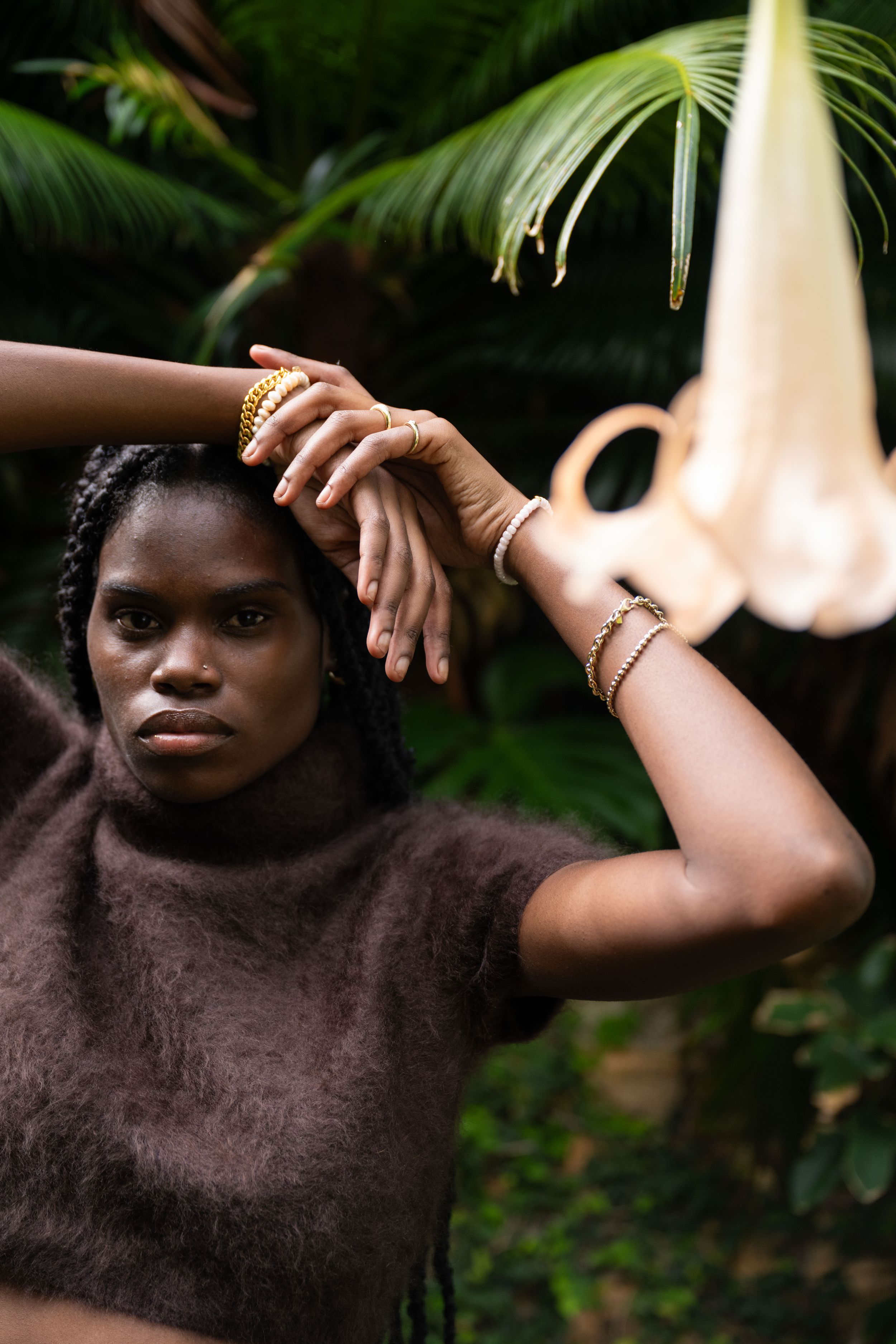 Fashion portrait with brown sweater and delicate gold jewelry against tropical palm fronds in natural lighting.