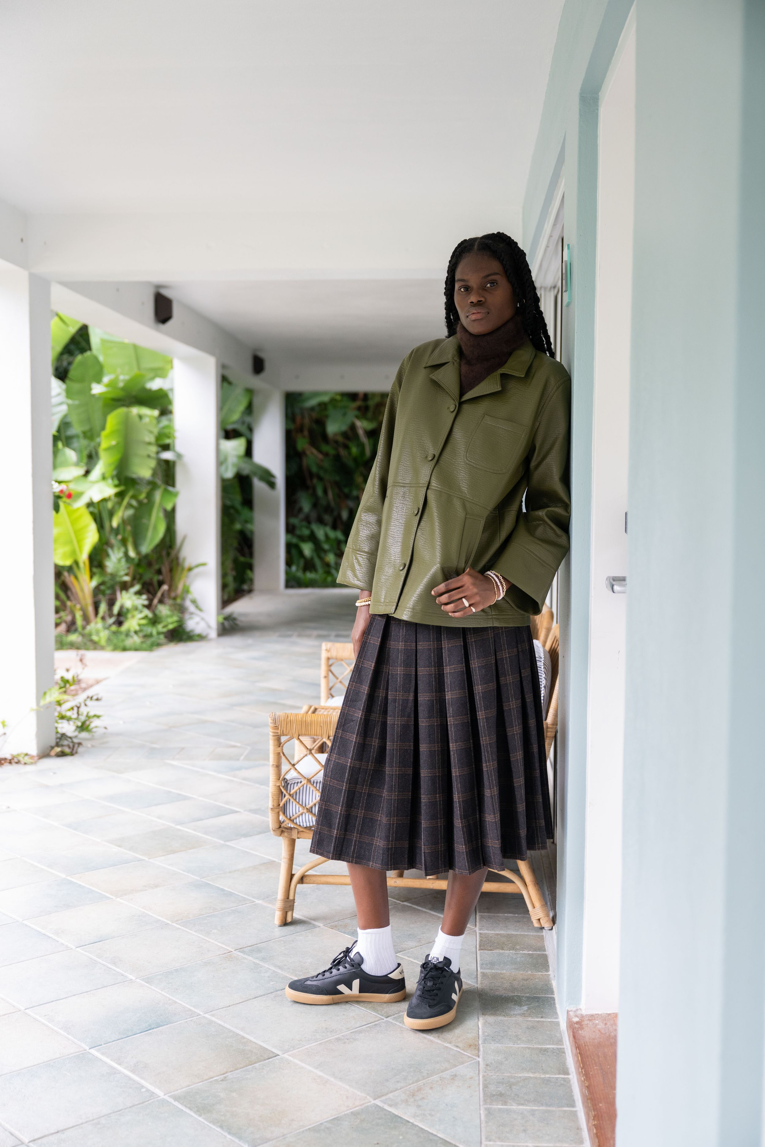 A person wearing a pleated gray skirt and olive green jacket stands in a bright white porch area with plants visible.
