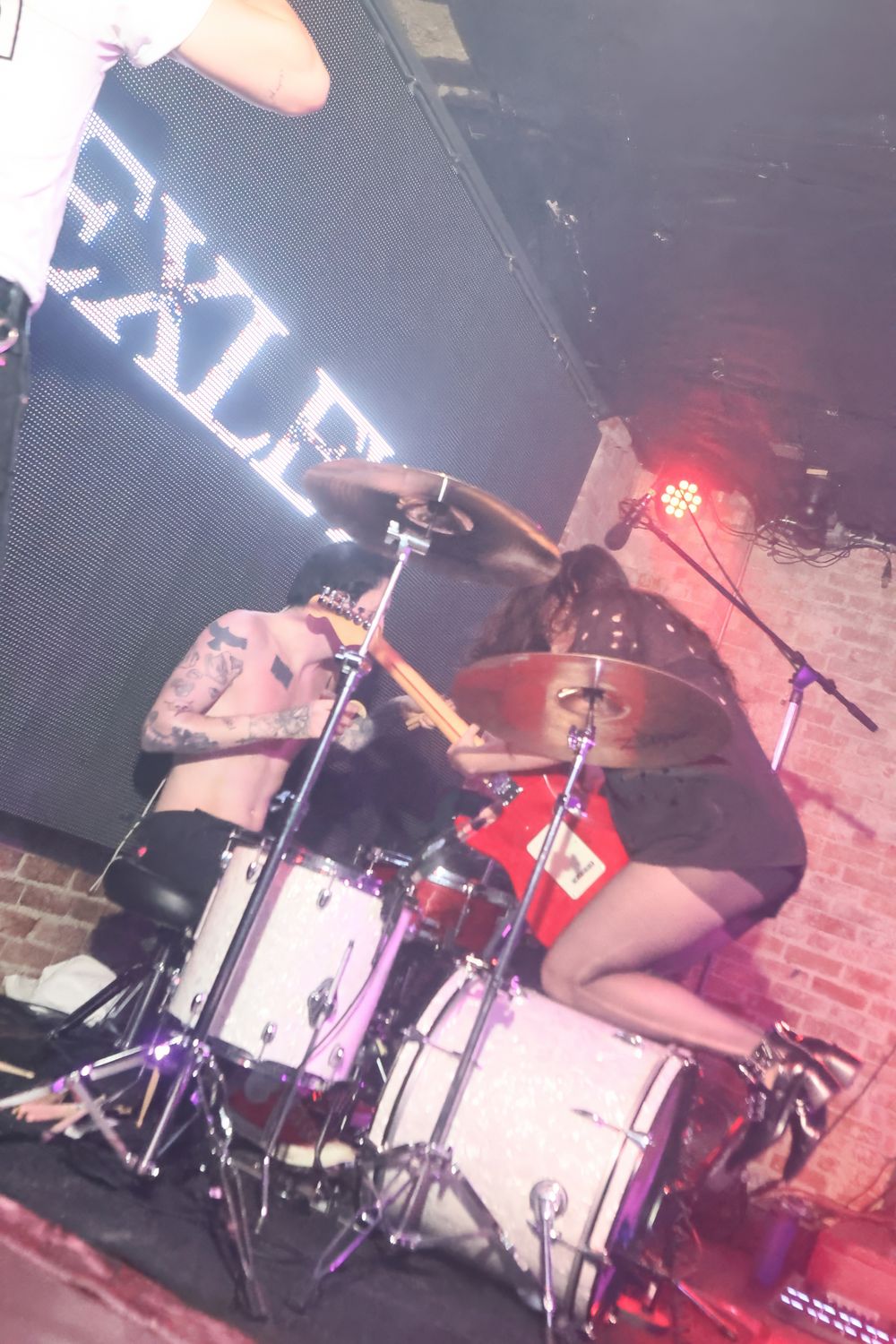 Drummer performs energetically on stage with drum kit under moody red lighting at a live music venue.