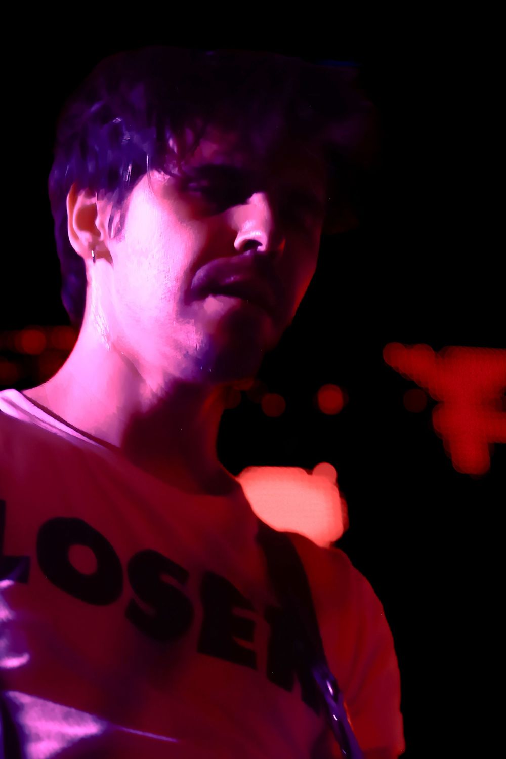 A person in a t-shirt illuminated by purple and red stage lighting against a dark background.