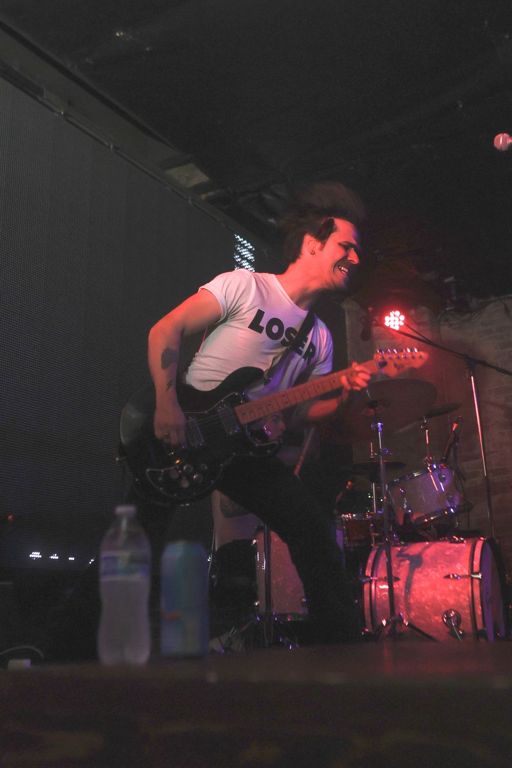 Musicians perform energetically on a dimly lit stage with red lighting and drum kit visible in the background.