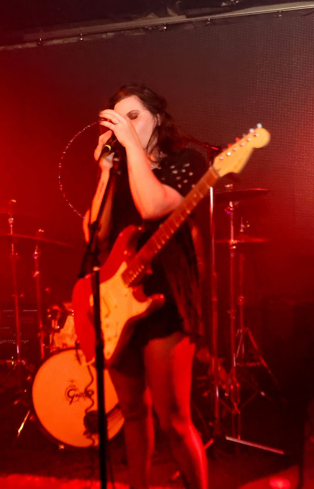 A musician performs on stage with a guitar under dramatic red lighting while a drum kit is visible in the background.
