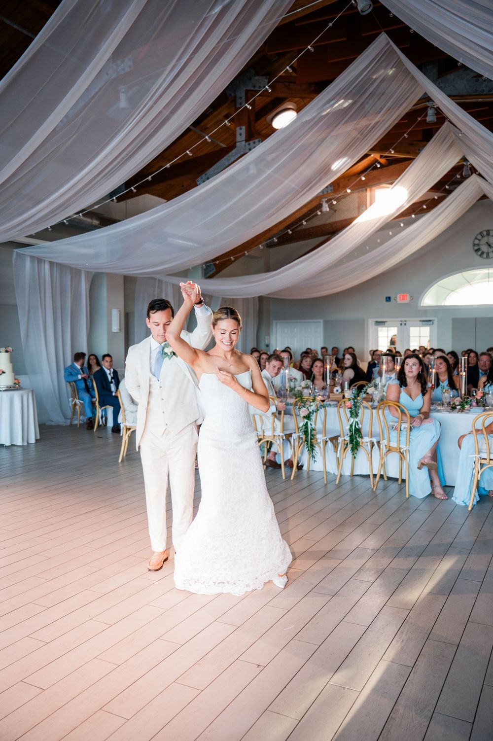 Wedding guests dance and celebrate on a light wooden floor under elegant white draped ceiling decorations at a reception.