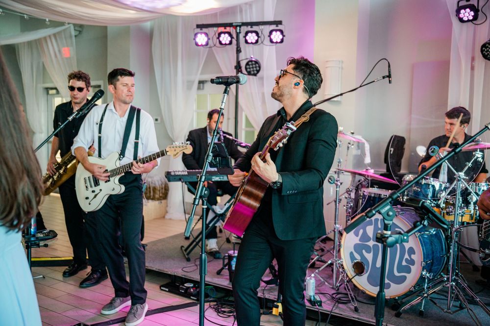 Musicians perform on stage with guitars and drums in a pink-lit venue during a live concert performance.