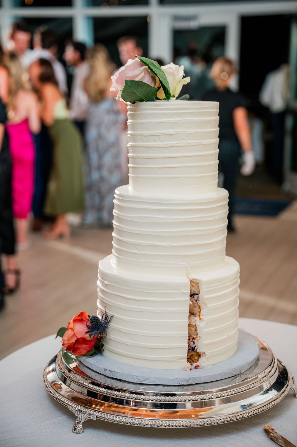 Elegant three-tiered white wedding cake with horizontal ribbed texture and floral decorations on silver platter at reception.