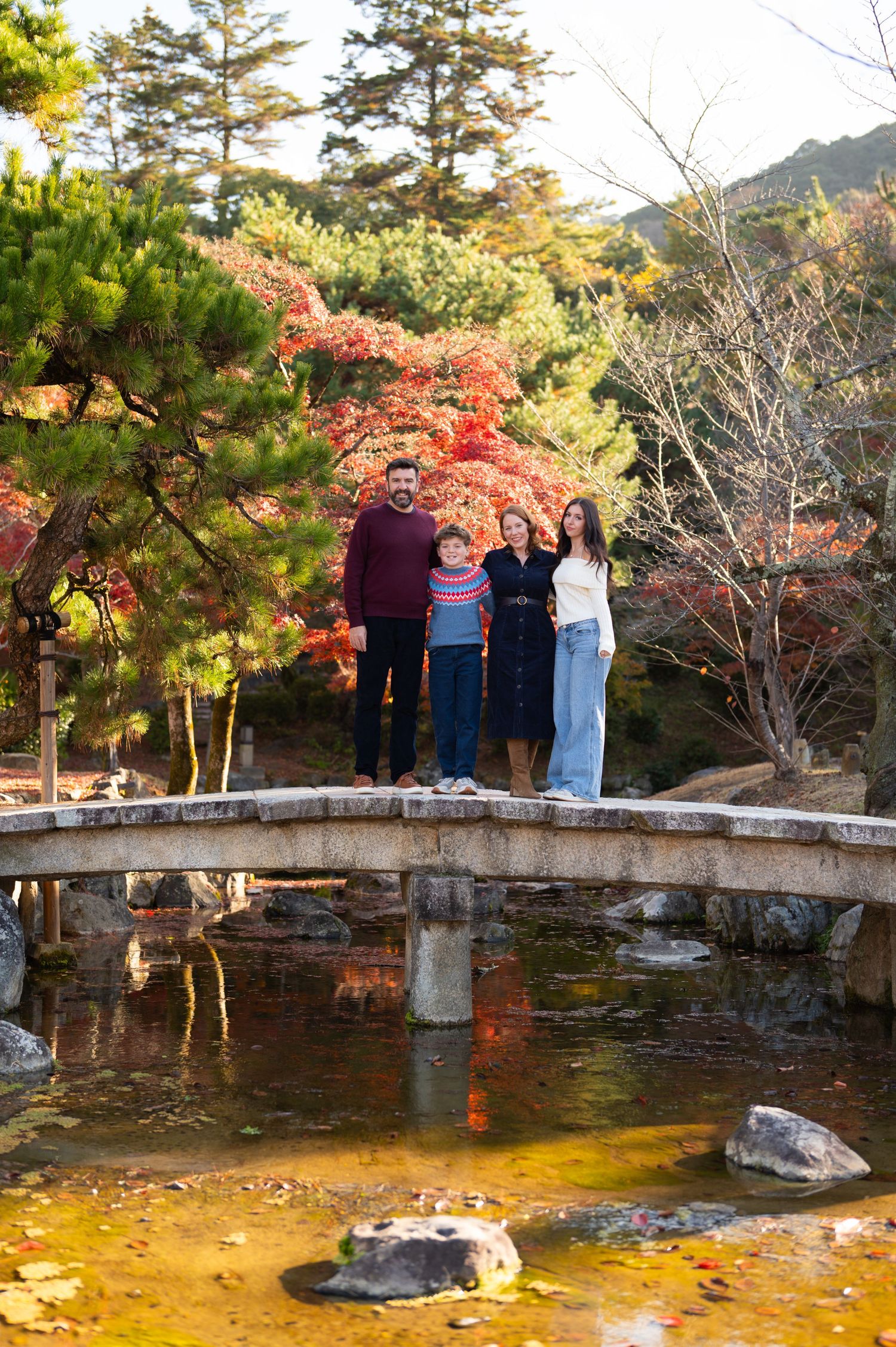 Family standing on an arched stone bridge surrounded by vivid autumn foliage in Japan.