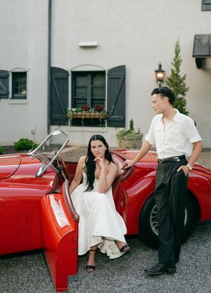 A couple poses casually against a vintage red convertible car in front of a gray building.